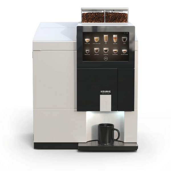 A Keurig commercial coffee machine with whole bean hoppers on top and a digital touchscreen panel showing various drink options. A black mug sits on the drip tray beneath the dispenser light.
